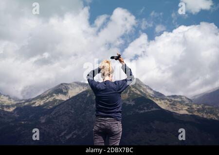 Rückansicht Porträt der kaukasischen blonden Frau Anordnung ihrer Haare binden in einem Brötchen vor einer Bergkette mit hohen Gipfeln und blauen Himmel mit dramatischen Stockfoto