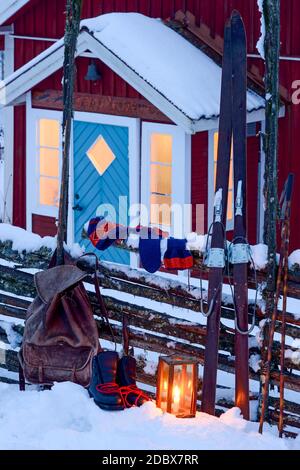 Eine Laterne, ein Rucksack, ein Paar Boote, Fäustlinge und ein Paar Himmel auf einem Zaun vor einer Veranda zu einem traditionellen schwedischen roten Haus im Winter lehnt Stockfoto