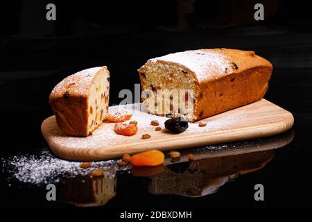 Süßer Cupcake in Form von Brot. Cupcake mit Rosinen, getrockneten Aprikosen, Pflaumen. Brot - Cupcake auf einem Holzbrett auf schwarzem Hintergrund, bestreut w Stockfoto