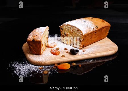 Süßer Cupcake in Form von Brot. Cupcake mit Rosinen, getrockneten Aprikosen, Pflaumen. Brot - Cupcake auf einem Holzbrett auf schwarzem Hintergrund, bestreut w Stockfoto