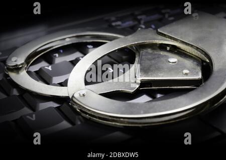 Ein konzeptuelles Bild mit Stahlhandschellen für Strafverfolgungsbehörden, die auf einem dunklen Tastenfeld für Online-Rechtssysteme angebracht sind. Stockfoto