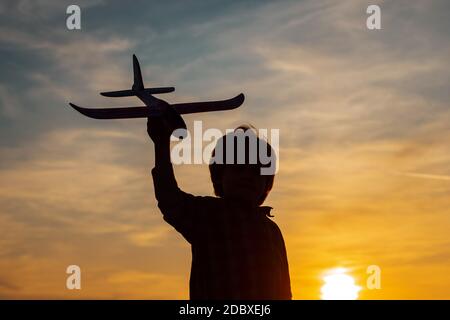 Kleiner Junge Flieger über Sonnenuntergang. Kleine Kinder mit Spielzeug Flugzeug in einem Feld bei Sonnenuntergang. Erfolg und Kind Leader Konzept. Stockfoto