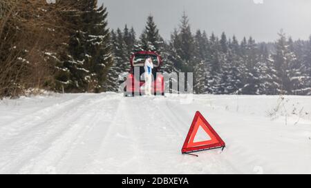 Leuchtend rote Dreieck auf einer verschneiten Straße eine Warnung an andere Fahrer mit Frau, die neben dem kaputten Auto im Wald, Reparatur Service auf Ihrem Mobiltelefon Stockfoto