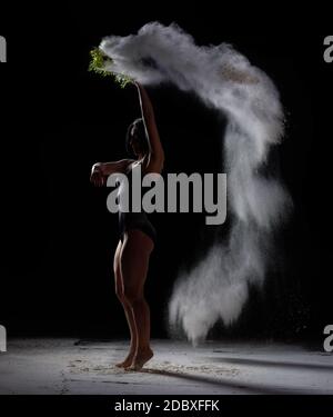 Junge schöne Frau mit kurzen schwarzen Haaren tanzt auf einem dunklen Hintergrund und hält ein Sieb mit Mehl. Sportlerin in schwarzem Bodysuit Stockfoto