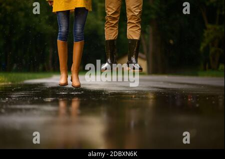 Liebespaar springt in einer Pfütze im Park, Sommer regnerischen Tag. Mann und Frau unter Regenschirm im Regen, romantisches Datum auf dem Fußweg, nasses Wetter in der Gasse Stockfoto