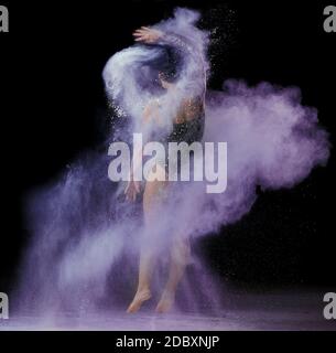 Schöne kaukasische Frau in einem schwarzen Body mit einem Sport Figur tanzt in einer purpurnen Mehlwolke Schwarzer Hintergrund Stockfoto