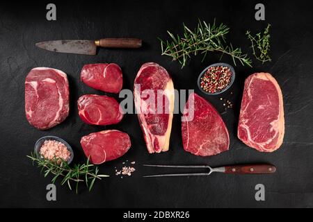 Fleisch, verschiedene Schnitte, darüber liegende flache Lay Shot mit einem Messer, eine Grillgabel, Salz, Pfeffer und Rosmarin auf dunklem Hintergrund Stockfoto