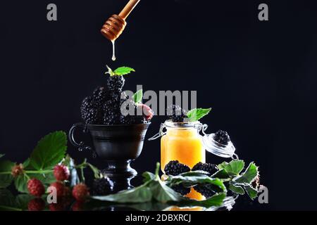 Ein Tropfen Honig aus einem Holzstock fällt auf ein BlackBerry. Beeren in einem Black Metal Vintage Cup mit Minzblättern. Speicherplatz kopieren. Glas mit Honig. Stockfoto