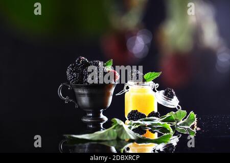 Brombeeren in einem schwarzen Metallbecher und einem Glas Honig auf schwarzem Hintergrund. Speicherplatz kopieren. Das Konzept des Stilllebens. Sommersaison. Beeren aus dem Fokus Stockfoto