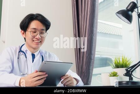 Glücklich asiatische junge Arzt Handsome Mann Lächeln Arbeit aus dem Home Office tragen Brille, mit schwarzen modernen smart digital Tablet-Computer auf dem Schreibtisch im Krankenhaus o Stockfoto