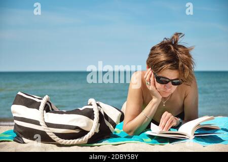 Nahaufnahme einer kaukasischen Frau mittleren Alters im Bikini, auf dem man sonnenbaden kann Der Strand und das Lesen eines Buches am hellen Sommertag Stockfoto