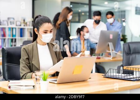Asiatische Büro Mitarbeiter Geschäftsfrau tragen Schutzmaske Arbeit in Neue normale Büro mit interracial Business-Team im Hintergrund als Soziale Distanc Stockfoto