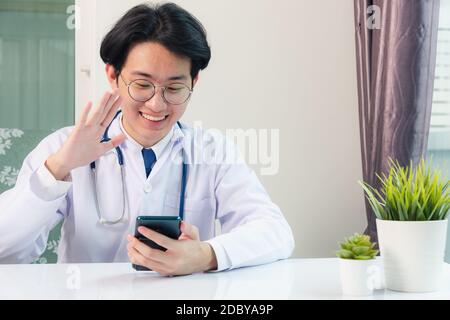 Asiatische Arzt junge gut aussehende Mann lächelnd Video-Anruf heben Sie die Hände, um Patienten mit digitalen Smart-Handy im Krankenhaus Schreibtisch Büro begrüßen, Technologie h Stockfoto