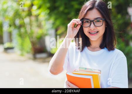 Portrait von Asian teen schöne junge Frau tragen Brillen Lächeln haben Zahnspangen auf Zähnen Lachen im Freien sie halten Bildung Bücher auf der Hand, M Stockfoto