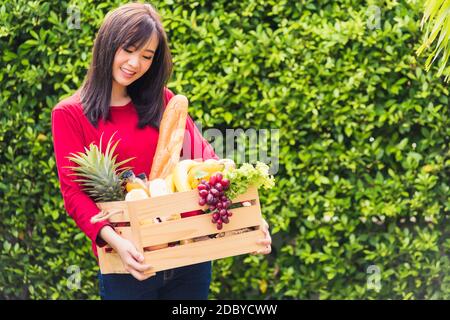 Porträt der asiatischen schöne junge Frau Bauer stehen sie lächeln und halten voll frische Lebensmittel rohes Gemüse Obst in einer Holzkiste in ihren Händen auf grün Stockfoto