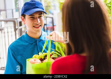 Asiatische junge Lieferung Mann in Uniform machen Lebensmittelgeschäft Service geben frisches Gemüse und Obst und Essen in grünen Stoffbeutel Frau Kunde an der Tür ho Stockfoto