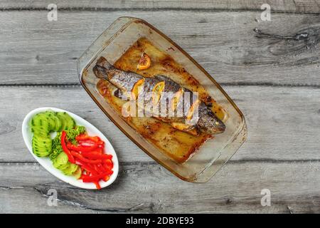 Table Top Blick auf gegrillte Forelle mit Zitronen in Glas Backform, und Schüssel von grüne Gurken, Zwiebeln und Paprika, auf grau Holzschreibtisch. Stockfoto