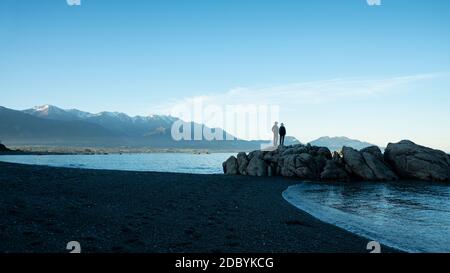 Pärchen stehen auf den Felsen am Strand mit schneebedeckten Bergen in der Ferne, Kaikoura, Neuseeland Stockfoto