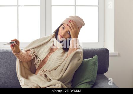 Frau mit einer hohen Temperatur in einem warmen Strickpullover und Hut hält ein Thermometer. Stockfoto