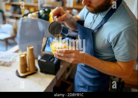 Der Barista in der Schürze gießt Schaum in den Kaffee im Café. Der Mann macht in der Cafeteria frischen Espresso, der Kellner an der Theke in der Bar Stockfoto