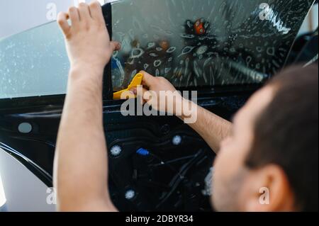 Männlicher Arbeiter installiert benetzte Auto Färbung, Tuning-Service. Mechaniker, der Vinylfarbe auf das Fahrzeugfenster in der Garage aufgibt, getöntes Glas Stockfoto