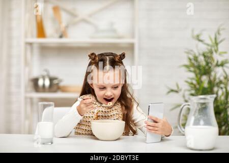 Nettes kleines Mädchen beim Frühstück: Müsli mit der Milch und Blick auf das Smartphone in der Küche. Gesundes Frühstück Stockfoto