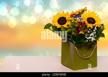 Nahaufnahme eines schönen Buketts mit gelben Sonnenblumen in einer dekorativen grünen Geschenkbox auf einem gelben Tisch auf einem verschwommenen blaugelben Hintergrund. Makro. S Stockfoto