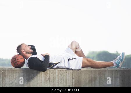 Der ruhende junge Basketballspieler liegt und entspannt sich mit Basketballball auf weißem Hintergrund. Stockfoto