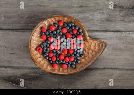 Table Top Aussicht auf eine gemischte Heidelbeeren und Himbeeren in Vintage geschnitzte hölzerne Platte auf grau Holz Schreibtisch platziert. Stockfoto