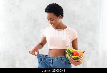 Glückliche afroamerikanische Frau in alten übergroßen Jeans halten Schüssel Mit Obst und Gemüse auf grauem Hintergrund Stockfoto