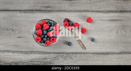 Table Top Aussicht auf kleine Glasschale mit Mix aus Heidelbeeren und Himbeeren gefüllt, silbernen Löffel auf der Seite, auf grau Holz Schreibtisch platziert. Stockfoto