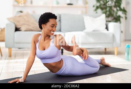 Sportliche afroamerikanische Frau, die ihr Bein auf einer Yogamatte streckt Zu Hause Stockfoto