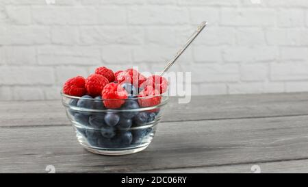 Kleines Glas Schüssel voll von Blaubeeren mit Himbeeren auf die Oberseite, und silbernen Löffel, auf grauen Schreibtisch aus Holz mit weißen Wand im Hintergrund platziert. Stockfoto