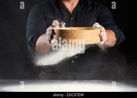 Ein Mann in schwarzer Uniform hält ein rundes Holzsieb In seinen Händen und siebt Weizenmehl auf einem Schwarzer Hintergrund Stockfoto