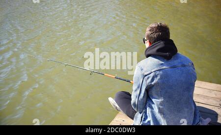 Junger Fischer in Jeansjacke sitzt auf hölzernen Pier, Angeln im See. Er hält die Rute und Blick auf Schwimmer. Stockfoto