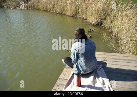 Junger Fischer in Jeansjacke sitzt auf hölzernen Pier, Angeln im See. Er hält die Rute und Blick auf Schwimmer. Stockfoto