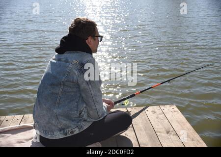 Junger Fischer in Jeansjacke sitzt auf hölzernen Pier, Angeln im See. Er hält die Rute und Blick auf Schwimmer. Stockfoto