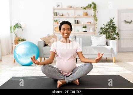 Schwarze schwangere Dame, die in Lotushaltung auf einer Yogamatte sitzt, meditiert oder Atemübungen macht und sich um ihre Gesundheit kümmert Stockfoto