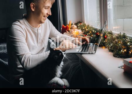 Junger Mann Freiberufler in Pyjama Arbeitsabstand online mit Videoanruf auf Laptop-Computer mit Katze zu Hause im Büro. Weihnachtsstimmung, Neujahr decorati Stockfoto