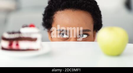 Afroamerikanische Frau, die zwischen Kuchen und Apfel wählt, guckt auf Lebensmittel von unter Tisch, Panorama Stockfoto