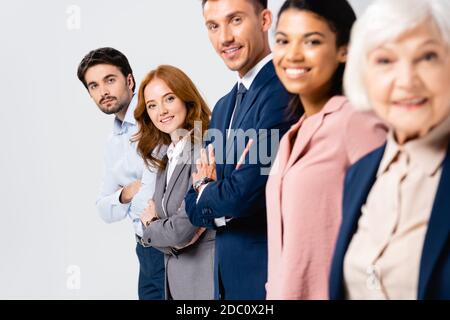 Lächelnde Geschäftsleute mit gekreuzten Armen blicken auf die Kamera in der Nähe von Multiethnic Kollegen auf verschwommenem Vordergrund isoliert auf grau Stockfoto