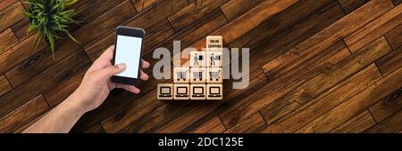 Hand mit Smartphone und Würfel mit Nachricht ONLINE-SHOPPING und Shopping-Symbole auf Holzhintergrund Stockfoto