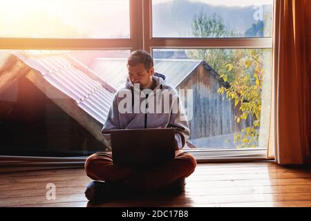 Mann mit Laptop sitzt auf dem Boden in der Nähe eines großen Fensters Stockfoto