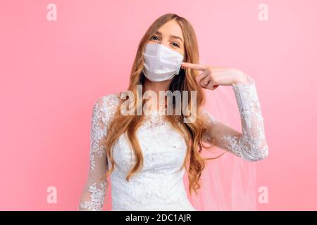 Eine Braut in einem Hochzeitskleid und eine medizinische Schutzmaske auf ihrem Gesicht zeigt auf die Maske auf einem rosa Hintergrund. Hochzeit, Quarantäne, Coronavirus Stockfoto