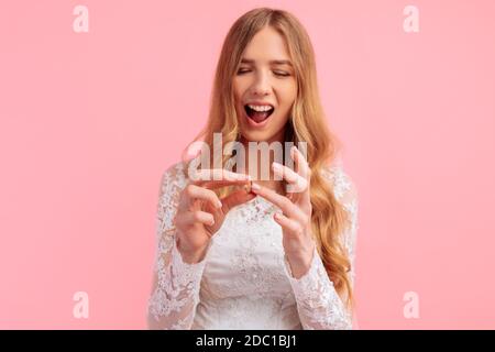 Eine fröhliche, aufgeregt Braut mit einem Blumenstrauß in einem Hochzeitskleid legt ihrem Finger einen Verlobungsring auf einem rosa Hintergrund auf. Hochzeit Stockfoto