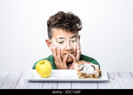 Ein Konzeptfoto eines Teenagers, der versucht, sich zwischen einem gesunden Apfel und einer süßen Zimtrolle zu entscheiden. Stockfoto