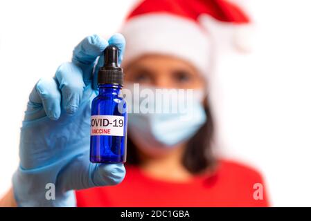 Covid19 Impfstoff weihnachtskonzept. Junge Frau mit Gesichtsmaske in santa claus Outfit hält covid Impfstoff in blauen Fläschchen und Spritze. Weiß backgrou Stockfoto