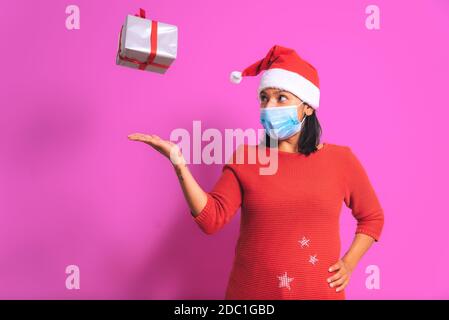 Covid-19 weihnachtskonzept. Glückliche Erwachsene Frau mit fliegenden Geschenk-Box, Gesichtsmaske, weihnachtsmann Outfit lächelnd auf rosa bunten Hintergrund. weihnachten durin Stockfoto