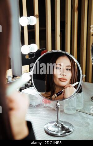 Spiegelung im runden Spiegel des Mädchens, das ihr Make-up macht. Das Mädchen hält einen Pinsel in den Händen. Stockfoto