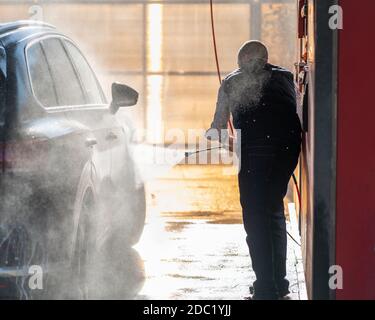 Der Mann wäscht das Auto bei der Autowäsche, gießt Wasser aus dem Schlauch. Sonnenlicht und Wasser spritzt in Kontur Stockfoto
